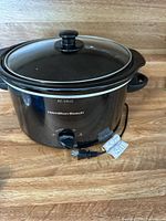 Black Hamilton Beach crock pot with glass lid, showing front and side views, with control knob and power cord visible.