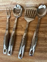 Four stainless steel Cuisinart utensils on wooden surface showing fork, slotted spoon, spatula, and serving spoon with logo on handles.