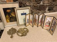Photo showing two brass trivets, 8-panel Japanese folding screen with painted figures, Burnes of Boston frame, and framed Bassi art print all displayed on a countertop.