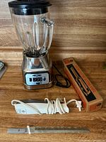 Full view of Oster blender with clear glass jar, stainless steel base with control panel showing multiple settings, black lid. GE electric cutting utensil and original box on wooden surface.