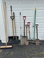 Full view showing all garden tools and black plastic garbage can lined against a garage door on driveway.