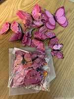 Large assortment of multiple pink butterflies on wooden surface, some loose and some in plastic packaging.