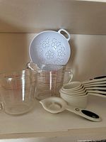 Photo showing large and small Pyrex glass measuring cups with red measurement markings, white plastic strainer sitting on top, and a set of white Oxo plastic measuring cups nested together.
