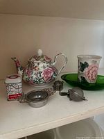 Wide view with floral rose teapot, London Tea tin, green relish tray, handleless floral patterned cup, and tea infusers visible.