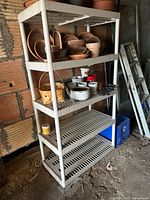 White plastic shelving unit with four slatted shelves. The upper shelves contain various terra cotta pots, plant saucers, ceramic pots, and baskets. The shelving unit is positioned against a brick wall with a metal ladder nearby.