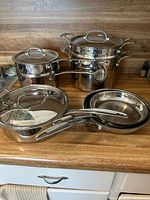 Full set of 10-piece Cuisinart stainless steel cookware on wooden countertop with all pieces visible including double boiler, frying pans, stock pot with insert, and saucepan.