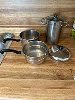Photo showing the double boiler set and asparagus pot with caged insert alongside their lids on a wooden kitchen countertop, highlighting their stainless steel finish and handles.