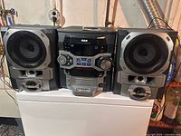 Front view of the entire RCA stereo shelf system including the main unit flanked by two speakers, all set on a white surface.