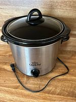 Rival Crock-Pot slow cooker with stainless steel body, black lid with handle, power cord visible on table surface.