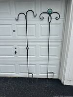 Full view of two different shepherd’s hooks leaning against a garage door. One with twisted metal shaft and two hooks, the other with curled arms and a glass orb.