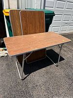 Three vintage folding tables with wood-look laminate tops and aluminum folding legs. One table is standing open; two tables are stacked folded behind it.