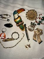 Photo showing assorted vintage jewelry items including carved toucan brooch, gold flower brooch, candy cane and ornament brooches, chains, rings, and pins on a light fabric background.