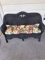 Front view of vintage black wicker settee sofa with floral seat cushion on porch carpet.