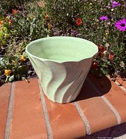 Celery green Bauer pottery planter showing swirl design and shape placed outdoors on brick surface with flowers in background.