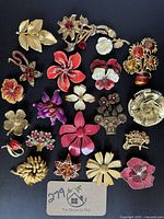 Full view of lot showing 21 vintage floral brooches in ivory, gold-tone, and cream hues with enamel, resin, and rhinestone details, arranged on black background