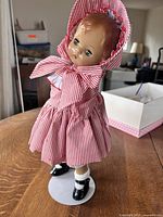 Full body image of the doll standing on display stand showing red and white striped dress, matching bonnet, black shoes, and socks.