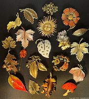 Top-down view of all 20 vintage fall-themed brooches arranged on black background showing a variety of leaves, a sun face, flower, and mushroom designs in enamel and metal tones.