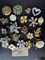 Top view of 20 vintage and retro floral brooches on black background showing enamel, rhinestone, and gold-tone finishes.