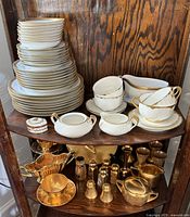 Top shelf of wooden étagère displaying white porcelain dinnerware with gold trim including plates, bowls, cups, saucers, creamers, sugar and serving pieces.