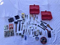 Full display of over 50 hand tools and hardware items including storage cases, bits, screwdrivers, hammers, pliers, scrapers, fasteners, measuring tools, tapes, and miscellaneous items spread out on white fabric background.