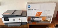 HP OfficeJet Pro 8025e printer with its original box placed on a table. The printer is light gray and dark gray with a touchscreen on the control panel.
