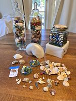 Two tall glass jars filled with mixed shells and marbles, one square glass jar with cork lid filled with marbles, large spiral shell, and various loose shells and marbles on wooden table