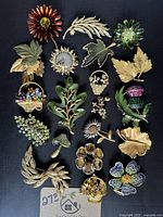 Photo showing the full collection of 24 vintage and modern brooches laid out on a dark background, featuring various leaf and flower designs with gold tone, enamel and rhinestone details.