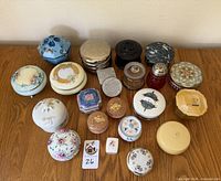 Full assortment of lidded vanity dishes, trinket boxes and powder containers displayed on a wooden surface, showing variety of sizes, materials and decorations.