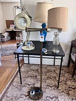 Four lamps on black table: white magnifying lamp, turquoise ceramic lamp with beige shade, cut crystal lamp with beige shade, and vintage brass swivel floor lamp in front.