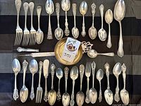 Overview of 30+ mixed sterling and silverplate flatware pieces arranged on black and gray cloth