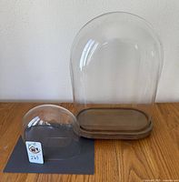 Photo showing two glass cloches, a large one with a wooden pedestal and a smaller dome without a base on a wood surface.