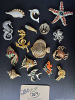 Front view of the 16 vintage nautical and sea life brooches on black background, showing varied designs including anchor, fish, starfish, shell, seahorse, crab, dolphin, octopus, and rope knot.
