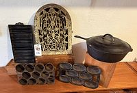 Overall photo showing vintage cast iron cookware including popover pan, cornbread pans, skillet with lid, and decorative heater vent.