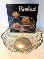 Silver plated wire basket with scalloped edges displayed in front of original black box with 'Basket' text and photo of baked goods inside basket.