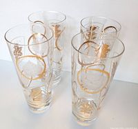 Six tall clear glass highball glasses with gold leaf and etched botanical leaf designs arranged in a group.