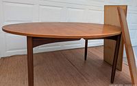 Full view of oval teak dining table with one extension leaf propped behind.
