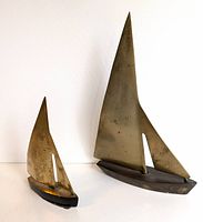 Front view of two vintage brass sailboat sculptures of different sizes, showing brass sails with cutout detail and wooden bases