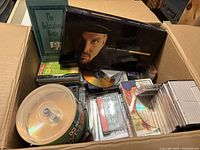 Overview of box containing mixed cassettes and CDs, showing boxed Garth Brooks cassettes, Relaxation Route cassette, loose tapes and stacked CDs.