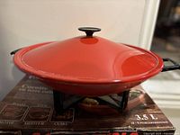 Red metal lid with black knob sitting on a black metal base with handles, showing 3.5 L capacity labeling and packaging beneath