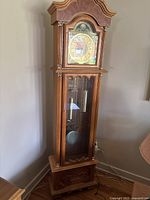 Full view of the tall light brown wooden grandfather clock standing in a corner. Shows bonnet top, glass door lower cabinet, and wood base with carvings.