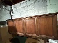 Front view of a wooden cedar chest in a dimly lit basement, showing paneled front with doors and sturdy legs.