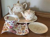 Photo of two vintage floral teapots on a white ceramic tray, a floral teacup on a square floral plate with matching spoon, and a small round plate with embossed text and floral pattern.