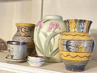 Photo shows three vases, one flower pot, and three small saucers arranged together on a shelf. The vases vary in color and design: one cream with pink flower relief, one yellow and grey with patterns, and one multicolored with scalloped rim. The flower pot is speckled white with floral decorations and paired with a saucer. The three additional small saucers are ceramic with scalloped edges and differing pastel colors.