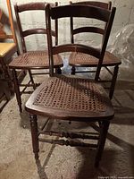 Three vintage wooden chairs with caned seats and ladder backs in an indoor setting on a concrete floor. One chair shows worn cane.