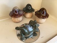 Three ceramic mushrooms with varying brown and green caps, and one ceramic frog figurine reclining on a small circular mirror base, displayed on a beige shelf corner.