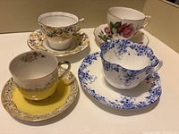 Four teacups placed on their saucers showing the variety of patterns and colors including yellow and gold, blue floral, pink rose, and gold floral motifs.
