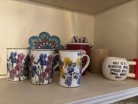Photo showing six assorted ceramic mugs and two floral decorative ceramic coasters on a shelf.