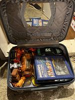 Top view of open black suitcase showing arrangement of vintage toy cars, plastic toys, and a Heroes & Heros tin box inside.