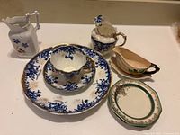 Overview of seven porcelain items including two blue and white floral teacups with saucers, large plate, small oval dish with floral detail and handle, small green and gold-rimmed plate, and two creamers with floral designs.