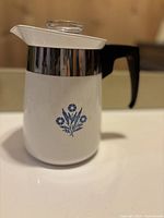 Front side view of the white Corning Ware percolator with blue cornflower design, black handle, chrome band, and white lid with glass knob.